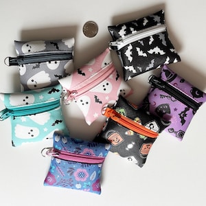 May include: Seven small square zippered pouches featuring various patterns including ghosts, bats, Halloween candy, and floral designs. Each pouch has a metal zipper and a small ring attachment. A single silver coin is placed near the top center.