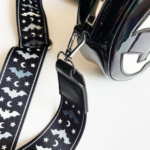 May include: Black, glossy handbag with a silver zipper and a detachable strap. The strap is black with a repeating pattern of bats, stars, and crescent moons. The bag has a white design on the front.