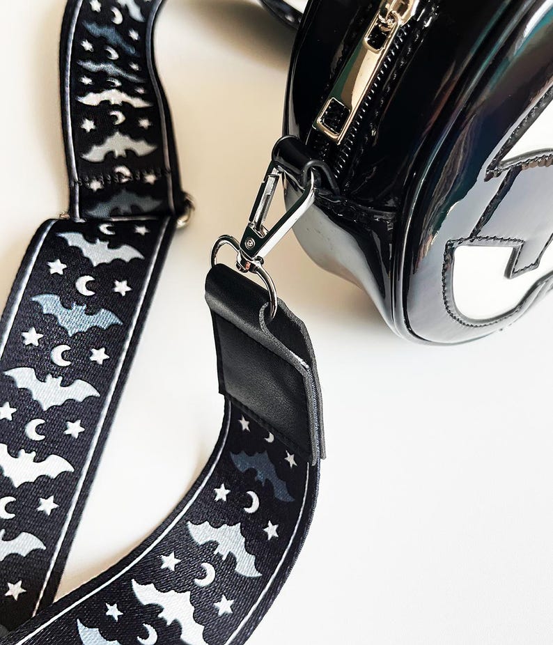 May include: Black, glossy handbag with a silver zipper and a detachable strap. The strap is black with a repeating pattern of bats, stars, and crescent moons. The bag has a white design on the front.