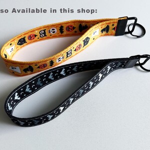 K&ouml;nnte beinhalten: Zwei Halloween-Armband-Schl&uuml;sselanh&auml;nger. Einer ist gelb mit orangefarbenen K&uuml;rbissen, schwarzen Katzen und wei&szlig;en Geistern. Der andere ist schwarz mit wei&szlig;en Flederm&auml;usen und Monden. Beide haben schwarze Metallbeschl&auml;ge.