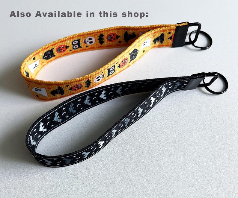 K&ouml;nnte beinhalten: Zwei Halloween-Armband-Schl&uuml;sselanh&auml;nger. Einer ist gelb mit orangefarbenen K&uuml;rbissen, schwarzen Katzen und wei&szlig;en Geistern. Der andere ist schwarz mit wei&szlig;en Flederm&auml;usen und Monden. Beide haben schwarze Metallbeschl&auml;ge.