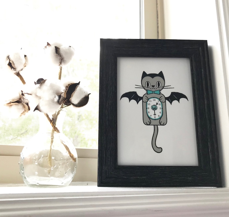 May include: A framed print featuring a cartoon cat with bat wings holding a clock. The cat is gray with a teal bow tie. The print is in a black frame, and a vase of cotton stems sits nearby.