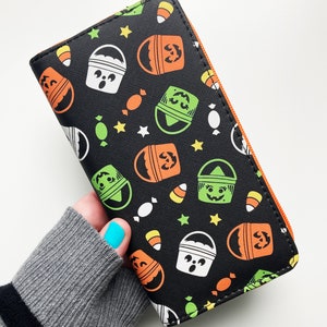 May include: Black wallet with a Halloween theme. The wallet has a repeating pattern of orange, green, and white buckets with jack-o-lantern faces, candy corn, and stars.