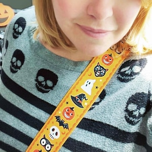 K&ouml;nnte beinhalten: Ein grauer Pullover mit schwarzen horizontalen Streifen und schwarzen Totenkopf-Designs. Ein gelber Gurt mit Halloween-Illustrationen, darunter K&uuml;rbisse, Flederm&auml;use und Geister, ist sichtbar.