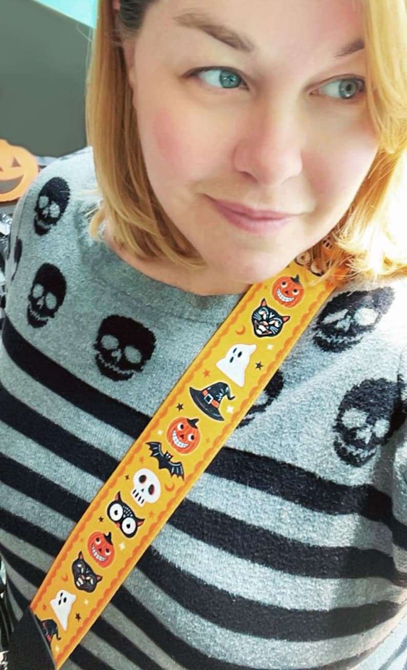 K&ouml;nnte beinhalten: Ein grauer Pullover mit schwarzen horizontalen Streifen und schwarzen Totenkopf-Designs. Ein gelber Gurt mit Halloween-Illustrationen, darunter K&uuml;rbisse, Flederm&auml;use und Geister, ist sichtbar.