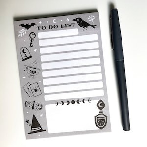 May include: A gray "TO DO LIST" notepad with black illustrations of a bat, raven, key, and potion bottle. A black pen rests beside the notepad. The notepad has a witchy, Halloween theme.