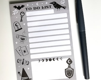 May include: A gray "TO DO LIST" notepad with black illustrations of a bat, raven, key, and potion bottle. A black pen rests beside the notepad. The notepad has a witchy, Halloween theme.