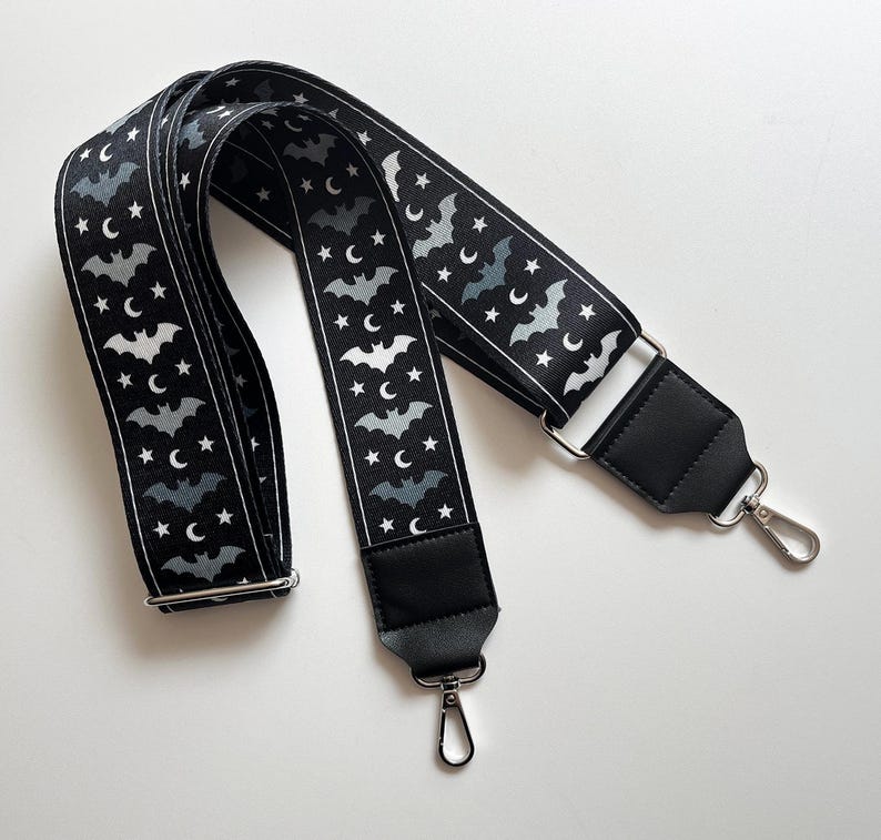 May include: Black and white patterned bag strap featuring a repeating design of bats, stars, and crescent moons. The strap has black leather ends and silver-tone metal hardware, including adjustable buckles and swivel clasps.