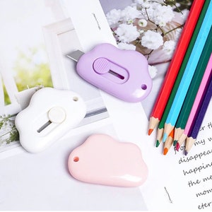 Puede incluir: Tres cúteres en forma de nube en blanco, lila y rosa. Una fila de lápices de colores rojos, verdes, azules y morados están dispuestos sobre una superficie blanca con texto impreso. Los cúteres tienen una cuchilla retráctil.