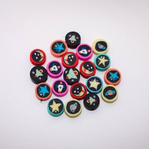 Puede incluir: Una colección de pequeñas cuentas redondas con temática espacial. Cada cuenta es negra, rosa o amarilla con ilustraciones coloridas de estrellas, cohetes, planetas y lunas. Las cuentas están dispersas sobre una superficie blanca.