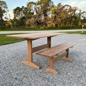 May include: A wooden table and bench set with a natural wood finish. The table has a rectangular top and a simple, sturdy base. The bench is also rectangular and has a similar base.
