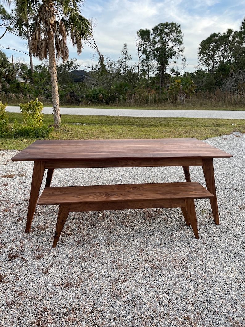 K&ouml;nnte beinhalten: Ein dunkelbraunes Holztisch- und Bankset im Mid-Century-Modern-Design. Tisch und Bank haben eine nat&uuml;rliche Holzoptik und abgeschr&auml;gte Beine.