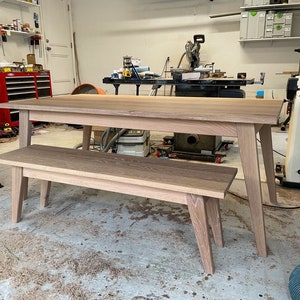 K&ouml;nnte beinhalten: Ein Esstisch- und B&auml;nkeset aus hellem Holz. Der Tisch hat eine rechteckige Platte und vier Beine. Die Bank hat eine rechteckige Platte und vier Beine. Das Set befindet sich in einer Werkstatt.