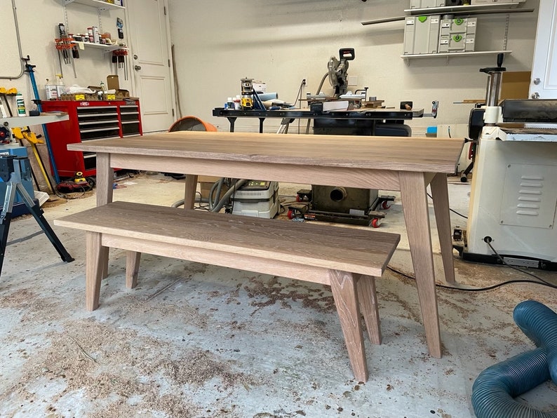 K&ouml;nnte beinhalten: Ein Esstisch- und B&auml;nkeset aus hellem Holz. Der Tisch hat eine rechteckige Platte und vier Beine. Die Bank hat eine rechteckige Platte und vier Beine. Das Set befindet sich in einer Werkstatt.