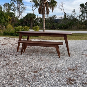 K&ouml;nnte beinhalten: Ein Esstisch- und B&auml;nkeset aus dunkelbraunem Holz. Der Tisch hat eine rechteckige Platte und vier Beine. Die Bank hat eine lange, rechteckige Sitzfl&auml;che und vier Beine. Das Set befindet sich im Freien auf einer Kiesfl&auml;che.