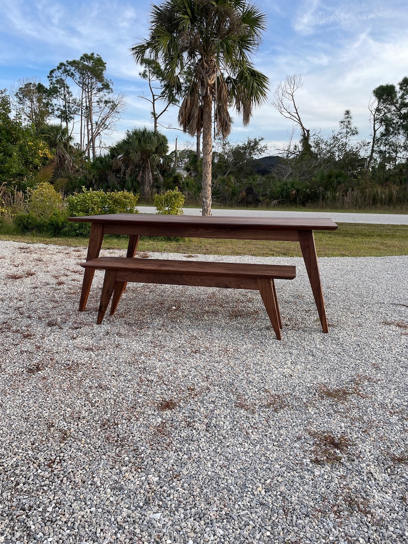 K&ouml;nnte beinhalten: Ein Esstisch- und B&auml;nkeset aus dunkelbraunem Holz. Der Tisch hat eine rechteckige Platte und vier Beine. Die Bank hat eine lange, rechteckige Sitzfl&auml;che und vier Beine. Das Set befindet sich im Freien auf einer Kiesfl&auml;che.