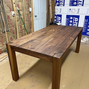 May include: A large, rectangular, dark brown wooden table with four legs. The table is made of solid wood with a natural grain pattern. The table is in a workshop setting.