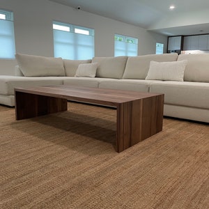 May include: A light brown wooden coffee table with a rectangular top and a wide, open base. The table is in front of a white sectional sofa with pillows.