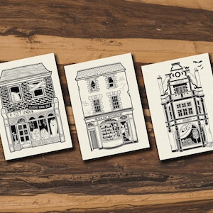 May include: Three black and white illustrations of storefronts. The first storefront says "6 Feet Under" and has a sign that says "Closing Down Sale". The second storefront says "Black Cat Apothecary". The third storefront says "Coffin & Co."