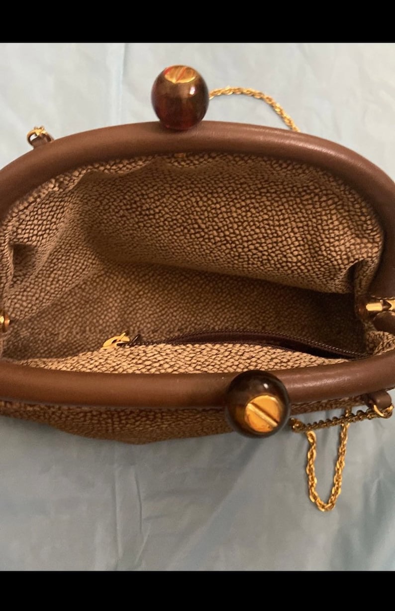 Puede incluir: Un bolso tejido marr&oacute;n con una correa de cadena dorada y dos cierres redondos marrones y dorados.
