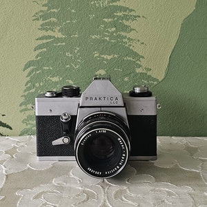 Op de afbeelding: Een zilveren en zwarte Praktica LLC camera met een zwarte lens op een wit kant tafelkleed voor een groene muur met een silhouet van bomen.