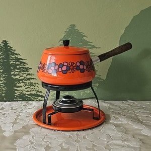 Op de afbeelding: Een oranje fonduepan met een zwarte standaard en een houten handvat. De pan heeft een bloemenpatroon in blauw- en roze tinten. De set bevat een ronde basis en een deksel met een zwarte knop. De achtergrond is een groene muur met geschilderde bomen.