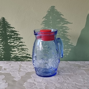 Peut inclure: Un pichet en verre bleu avec un couvercle rouge et une anse. Le pichet a une surface texturée et est décoré d'un motif floral.