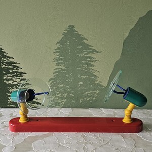 May include: A colorful toy with two clear fan blades attached to teal and yellow supports on a red base. The toy is set against a backdrop of a green wall with tree silhouettes and a white lace tablecloth.