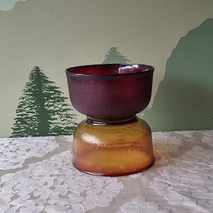 Op de afbeelding: Een tweekleurige glazen vaas met een donkerpaarse bovenkant en een licht amberkleurige onderkant. De vaas heeft een getextureerd oppervlak en staat op een wit tafelkleed.