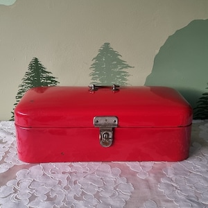 Op de afbeelding: Een rode metalen broodtrommel met een scharnierend deksel en een zilveren sluiting. De broodtrommel staat op een wit tafelkleed.