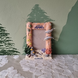 May include: A light pink and brown picture frame with a seashell design. The frame features a dolphin, fish, and seaweed. The text "Journey to Atlantis" is printed on the frame. The frame is sitting on a white tablecloth.