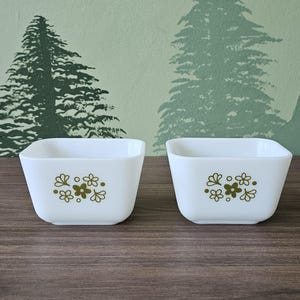 以下が含まれることがあります： 緑と黄色の花柄が施された白い正方形のセラミックボウルが2つ。ボウルは茶色の木製の表面の上に置かれています。