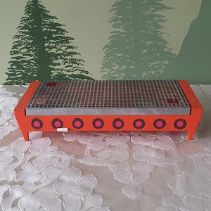 Op de afbeelding: Oranje elektrische voedselwarmer met een metalen grill bovenop en een rij roze en witte cirkels rond de basis.