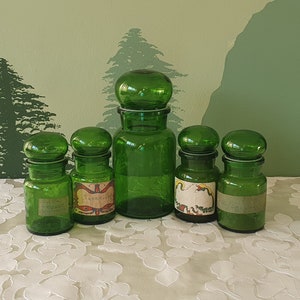 Może przedstawiać: Pięć zielonych szklanych słoików z etykietami i korkowymi korkami. Słoiki są ułożone w rzędzie na białym tiulowym obrusie.