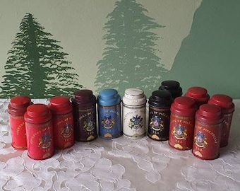 Może przedstawiać: Kolekcja 12 małych, okrągłych, metalowych puszek z różnymi wzorami. Puszki są ułożone w dwóch rzędach po sześć, przy czym górny rząd jest lekko przesunięty względem dolnego. Puszki są połączeniem czerwonego, niebieskiego i czarnego z złotymi akcentami. Puszki stoją na białym obrusie z kwiatowym wzorem.