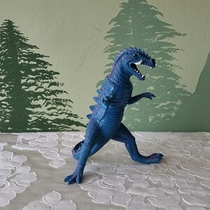 Op de afbeelding: Blauw plastic dinosaurus speelgoed met een stekelige rug en een wijd open mond. De dinosaurus staat op een wit tafelkleed met een bloemenpatroon.