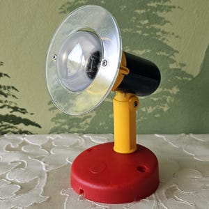 May include: A small, colourful spotlight with a red base, yellow stand, and black lamp housing. The lamp has a clear, circular lens and is designed to be adjustable. The background features a green wall with a tree pattern and a white lace tablecloth.