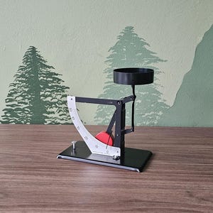 Op de afbeelding: Een zwart en zilver mechanisch apparaat met een rode bol, een gebogen witte schaal en een zwarte beker. Het apparaat staat op een zwarte basis, tegen een groen en wit geschilderde muur met boom silhouetten.