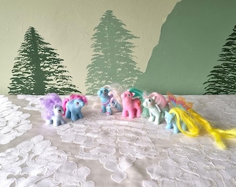 Könnte beinhalten: Eine Gruppe von acht pastellfarbenen Plüschponys mit langen, fließenden Mähnen und Schwänzen. Die Ponys sind in einer Reihe auf einer weißen, strukturierten Oberfläche angeordnet.