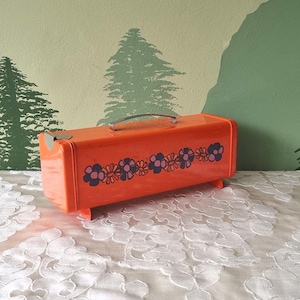 Op de afbeelding: Een oranje vintage reiskoffer met een zilveren handvat en bloemenpatroon. De rechthoekige koffer staat op kleine voetjes. Het bloemenontwerp is een herhalend patroon van paarse en blauwe bloemen.