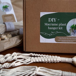 May include: A DIY macrame plant hanger kit in a brown cardboard box. The kit includes natural cotton cord, wooden beads, and a wooden ring. The box has a label that says "DIY Macrame plant hanger kit" and includes instructions on how to create the hanger.