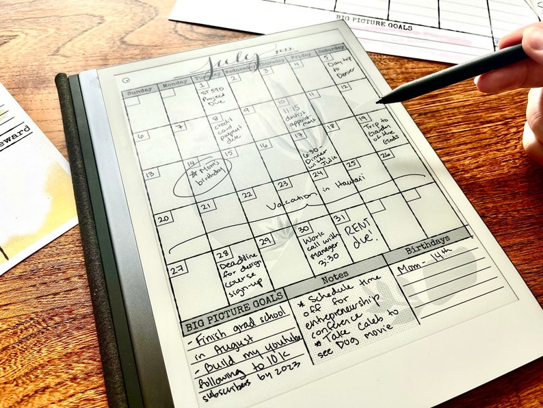 May include: A white digital calendar with a black frame and a black stylus. The calendar is open to July and has handwritten notes and appointments. The notes include "Mom's birthday", "Vacation in Hawaii", "Rent due", and "Schedule time off for entrepreneurship conference".