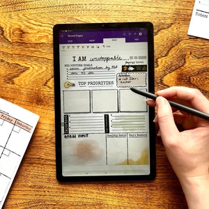 May include: A digital tablet with a white background and a handwritten to-do list. The list includes "I AM unstoppable", "BIG PICTURE GOALS", "earn promotion", "TOP PRIORITIES", "Important", "BRAIN VOMIT", "Healthy Habit", and "Day's Reward". The tablet is on a wooden surface and a person is using a stylus to write on the screen.