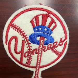 May include: A white felt patch with a red circle around the edge. Inside the circle is a red baseball bat with a blue and red baseball cap on top. The word "Yankees" is embroidered in red on the patch.