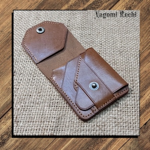 Puede incluir: Una cartera de cuero marrón con cierre de botón a presión, abierta para revelar el interior. La cartera está sobre una superficie texturizada, con el texto "Vagomi Rechi" visible. La cartera es de cuero liso marrón.