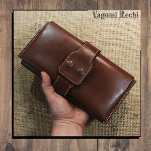 May include: A brown leather wallet with a flap closure secured by two brass-colored buttons. The wallet is held in a hand and features white stitching. The text "Vagomi Rechi" is visible in the upper right corner. The background is a wooden surface.