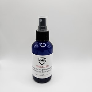 May include: A blue glass bottle with a black spray top and a white label. The label says "SHIELDED Keep the Negativity Away Protection Smudge Spray 4 oz."