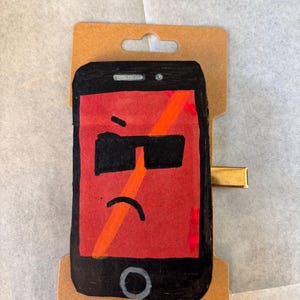 Può includere: Una molletta per capelli rettangolare con un'illustrazione disegnata a mano di uno smartphone nero con schermo rosso. Il telefono ha un'espressione imbronciata con occhiali da sole neri e una bocca curva. Una linea arancione diagonale attraversa lo schermo e una clip in metallo dorato è fissata sul lato del supporto in cartone marrone.