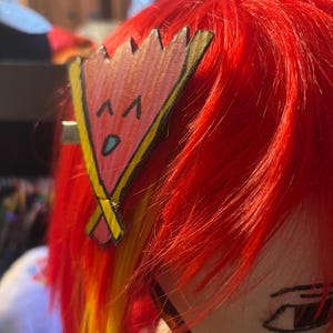 May include: A red wig with a yellow and orange streak. A pink and gold paper clip with a cartoon face is attached to the wig.