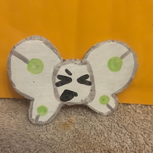 May include: Hand-painted, white butterfly-shaped artwork with a distressed, metallic silver outline. The butterfly has green polka dots and a black, angry face. The background is a solid orange color.